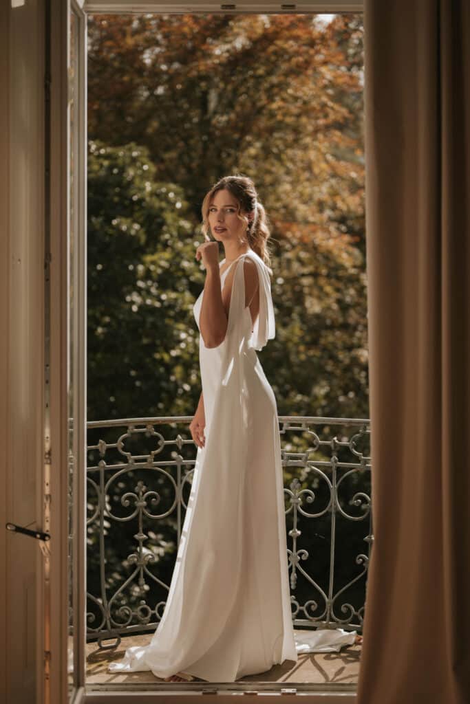 Mariée debout sur le balcon du manoir, entourée de verdure.