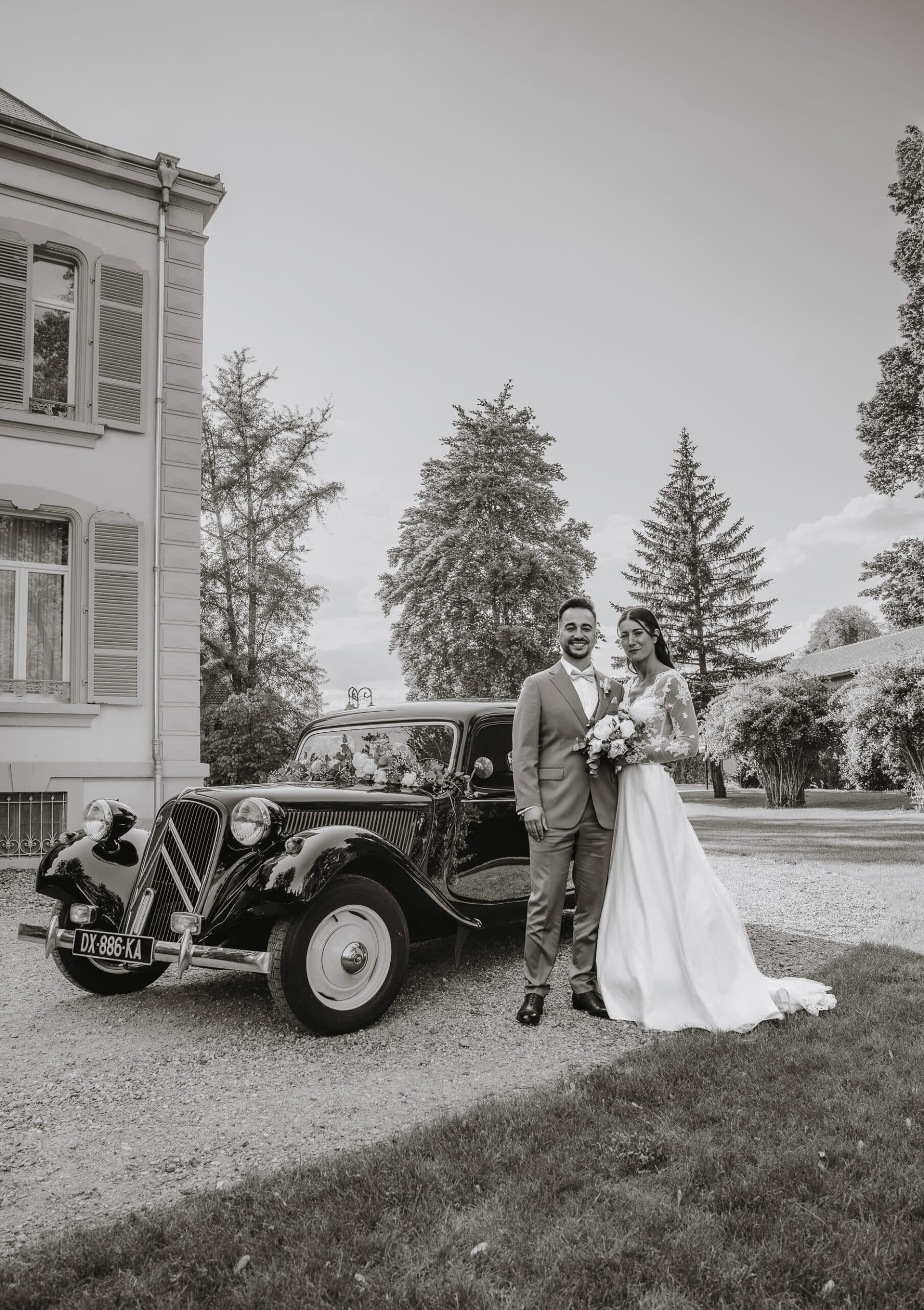 Couple de mariés posant devant une voiture de collection devant le domaine.