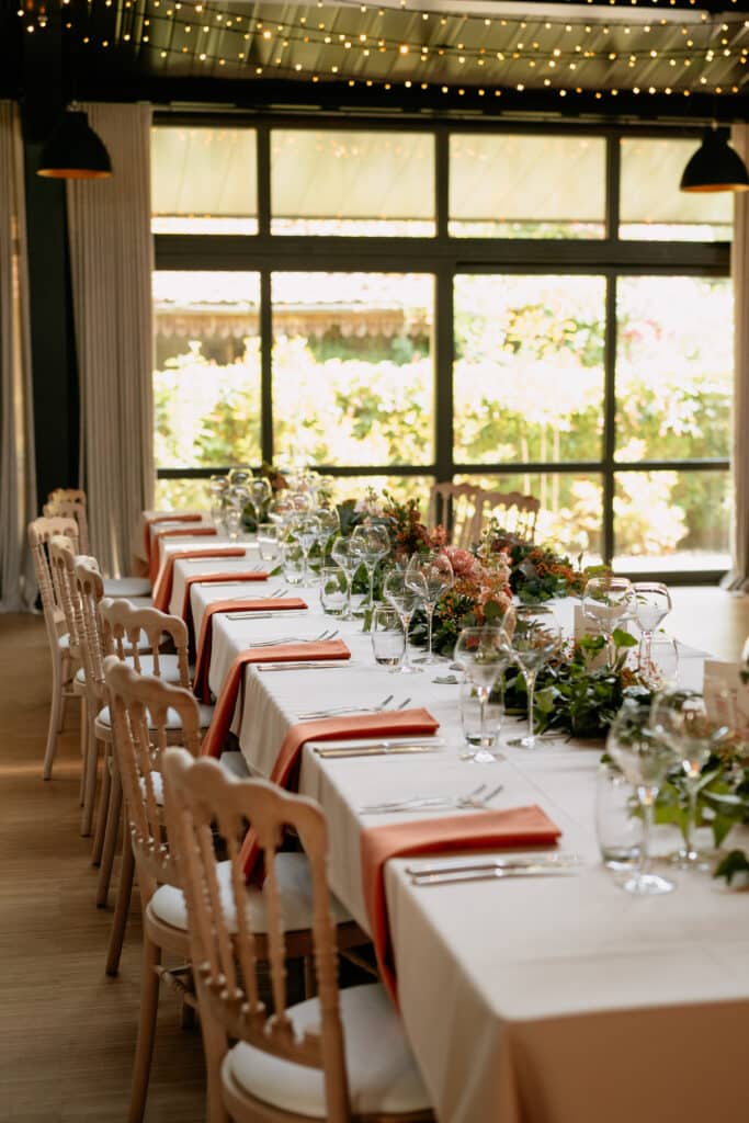 Table de réception dressée avec serviettes corail, verres élégants et décoration florale dans une salle lumineuse.