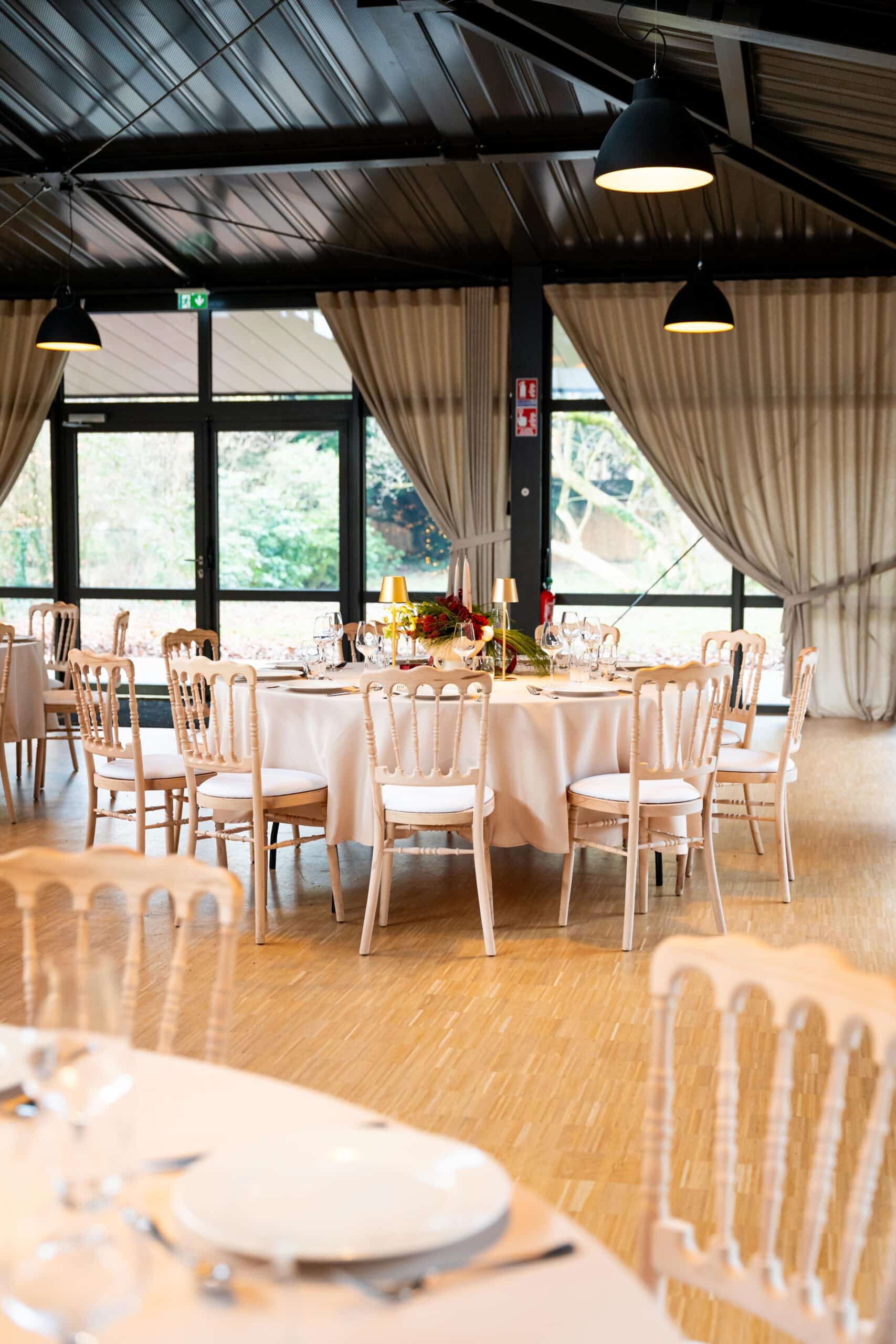 Round banquet table set for a formal event with white chairs, glassware, and a floral centerpiece in a bright venue with large windows and draped curtains.