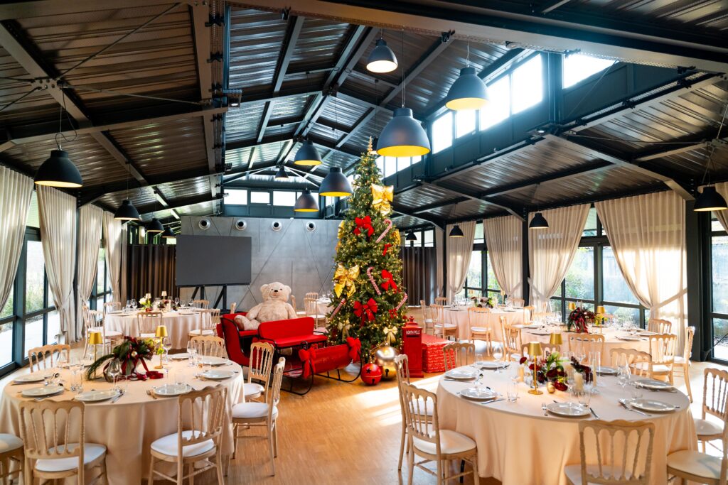 Repas de fin d'année – La villa des hêtres pourpres Festive event hall with a tall Christmas tree, gold ribbons, and a red sleigh centerpiece surrounded by round tables and beige chairs