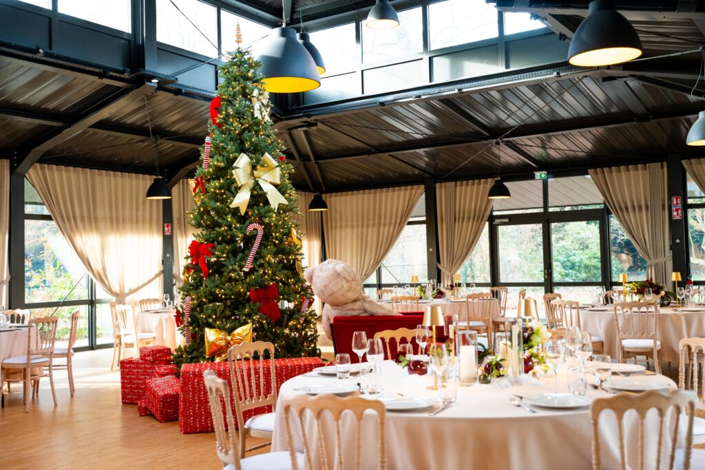 repas de fin d'année – La villa des hêtres pourpres Festive banquet hall with a tall Christmas tree, gold bows, candy canes, and wrapped presents beneath; a beige teddy bear sits on a red sofa nearby.