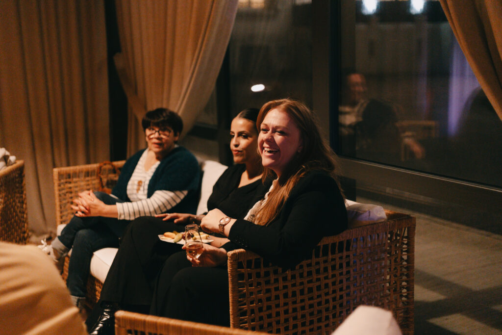 GalaBoRso-2026103 – La villa des hêtres pourpres Three women sitting on wicker chairs in a dim lounge; the woman in front-right laughs, holding a wine glass.