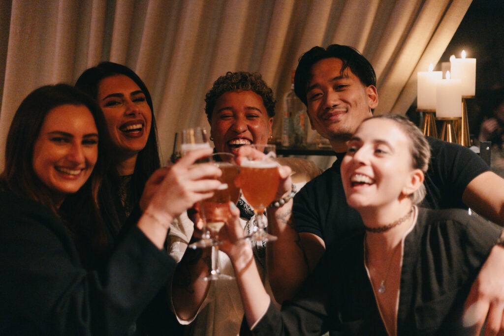 GalaBoRso-2026118 – La villa des hêtres pourpres Group of friends toasting with drinks, smiling in a warmly lit party setting.