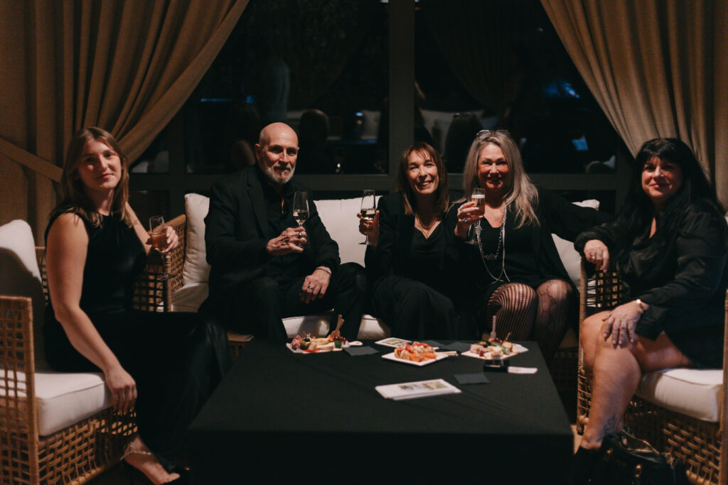 GalaBoRso-202617 – La villa des hêtres pourpres Group of five adults in black attire sitting on a sofa, holding drinks at a dim lounge with snacks on a table in front of them.