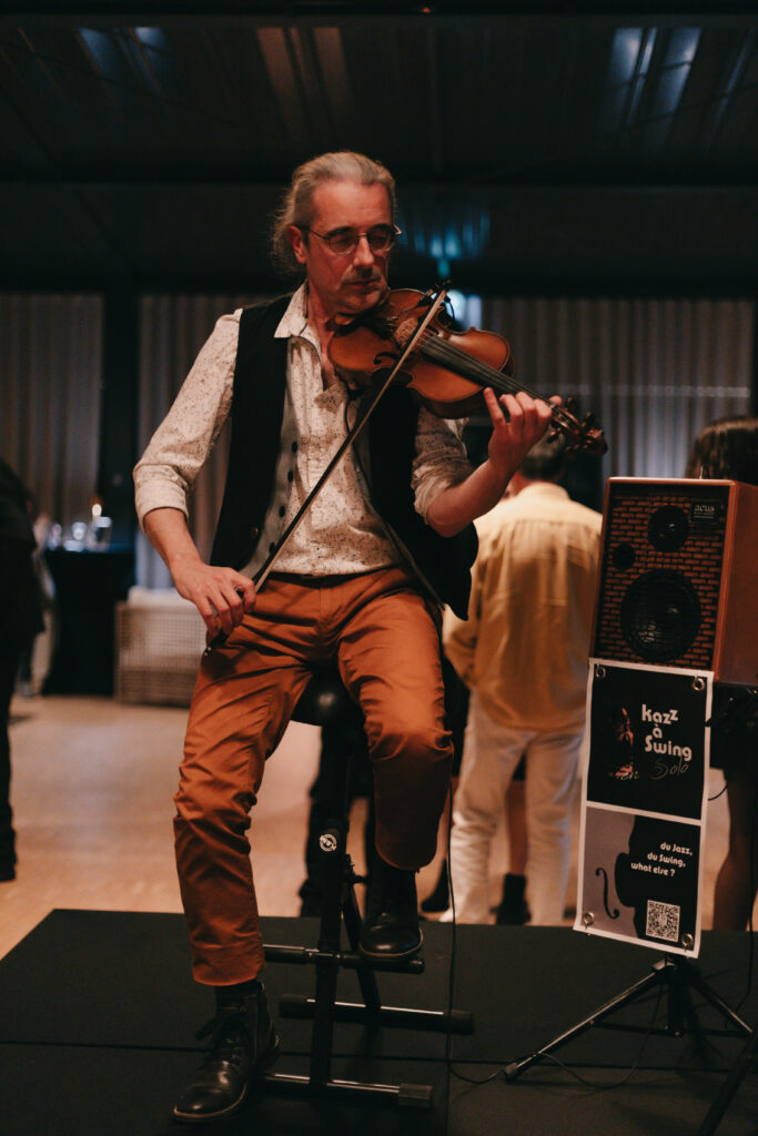 GalaBoRso-20262 – La villa des hêtres pourpres Male violinist performing live indoors, wearing glasses, a white patterned shirt and black vest while playing a violin on a small stage with a speaker nearby.