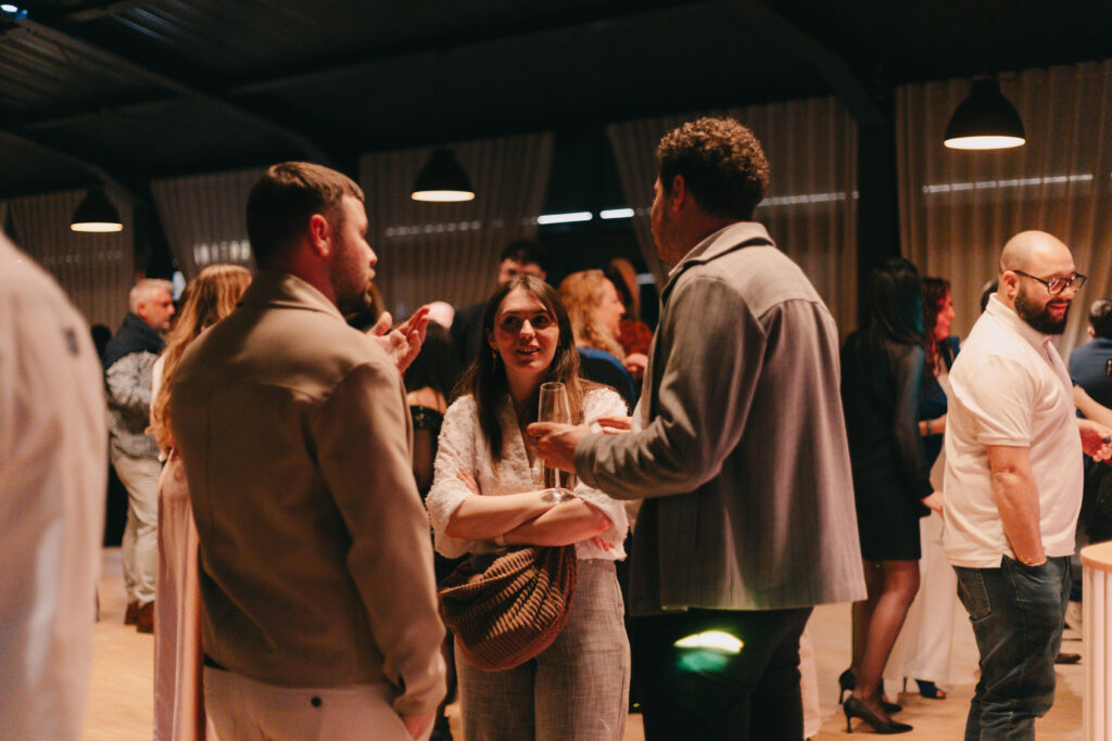 GalaBoRso-202628 – La villa des hêtres pourpres Woman in white top converses with two men at a crowded indoor social event, holding a wine glass in her hand.