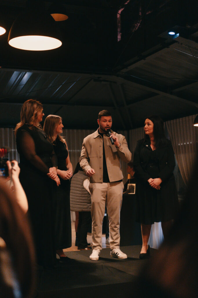 GalaBoRso-202652 – La villa des hêtres pourpres Man speaks into a handheld microphone on stage, flanked by four adults at a formal indoor event audience watching nearby