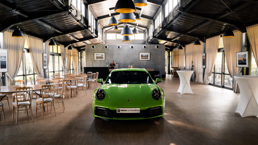 DSC06688 – La villa des hêtres pourpres Green sports car parked in the center of a bright event hall with wooden chairs, high ceilings, and hanging lamps.