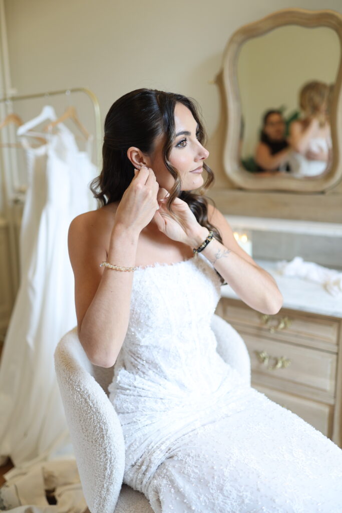 AM7A6076 – La villa des hêtres pourpres Bride in a lace wedding gown sits in a dressing room, placing earrings with a mirror in the background.