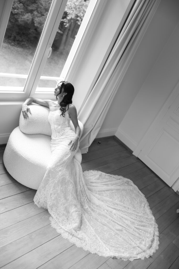 La robe de la Mariée – La villa des hêtres pourpres Bride in a lace wedding gown sits on a textured round chair by a large window, gazing to the side. (Black and white)