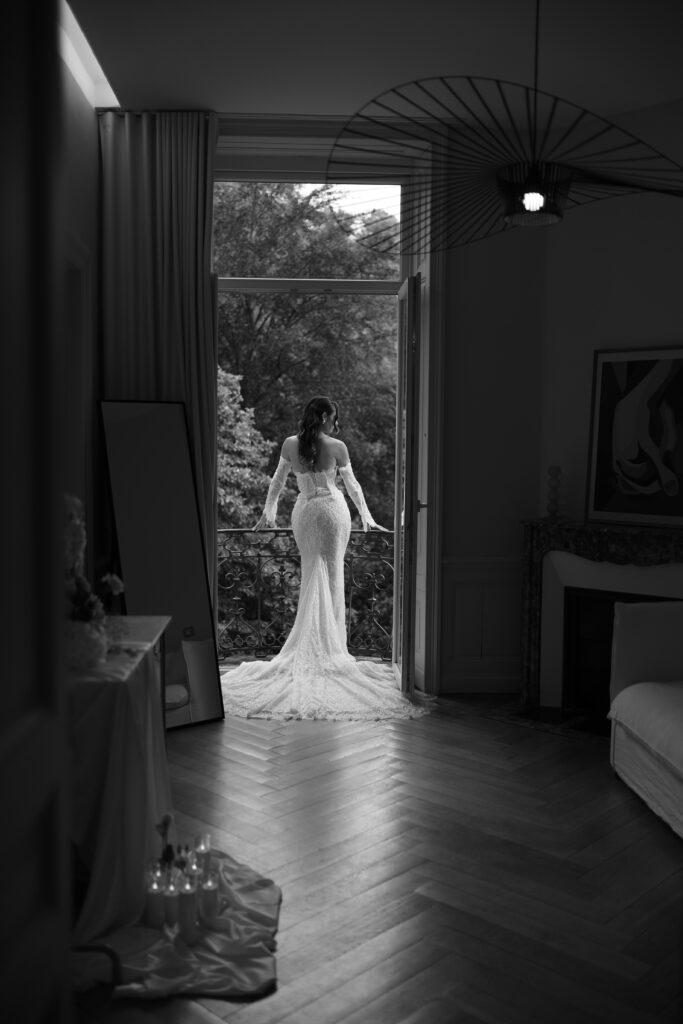 La mariée – La villa des hêtres pourpres Bride in a lace wedding gown stands at an open balcony door, back to us, lush trees outside, indoors with a chandelier overhead and wooden floor.