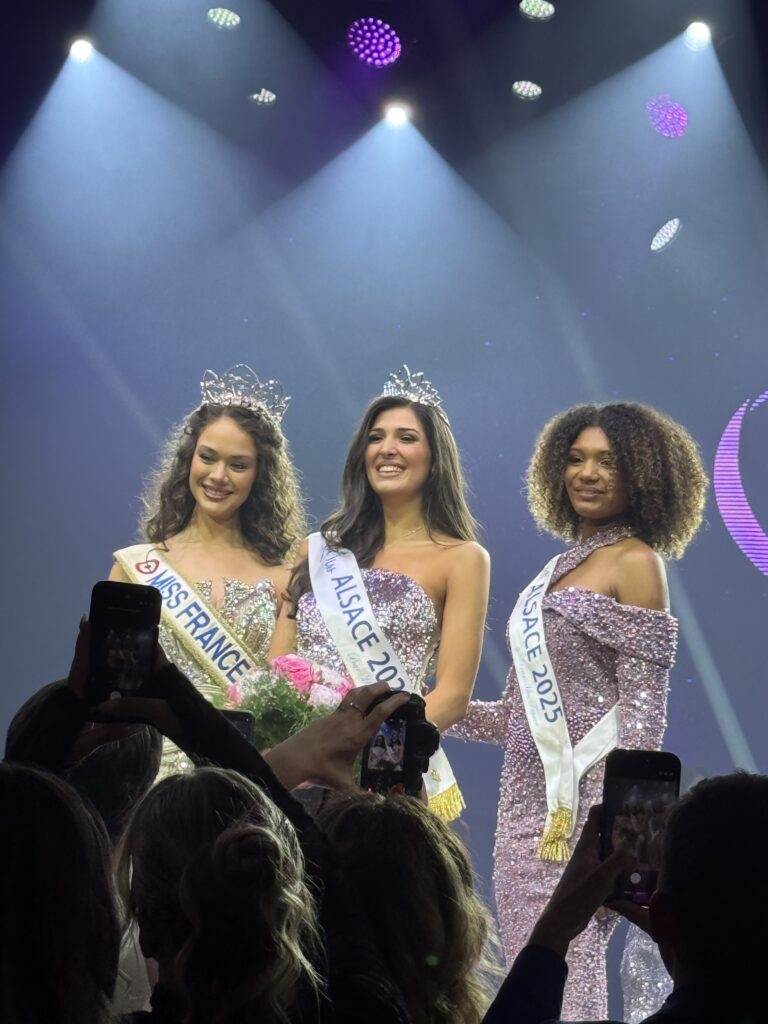 Courronnement de Miss Alsace 2026 – La villa des hêtres pourpres Three pageant winners wearing tiaras on stage, in sparkling gowns, posing for photos as audience members snap pictures.