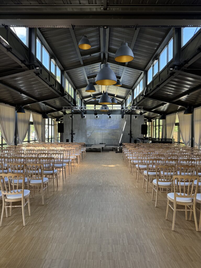 Conférence dans l'Orangerie – La villa des hêtres pourpres Industrial-style event hall with rows of wooden chairs facing a stage, blue-and-yellow pendant lamps overhead, and large side windows letting in light.