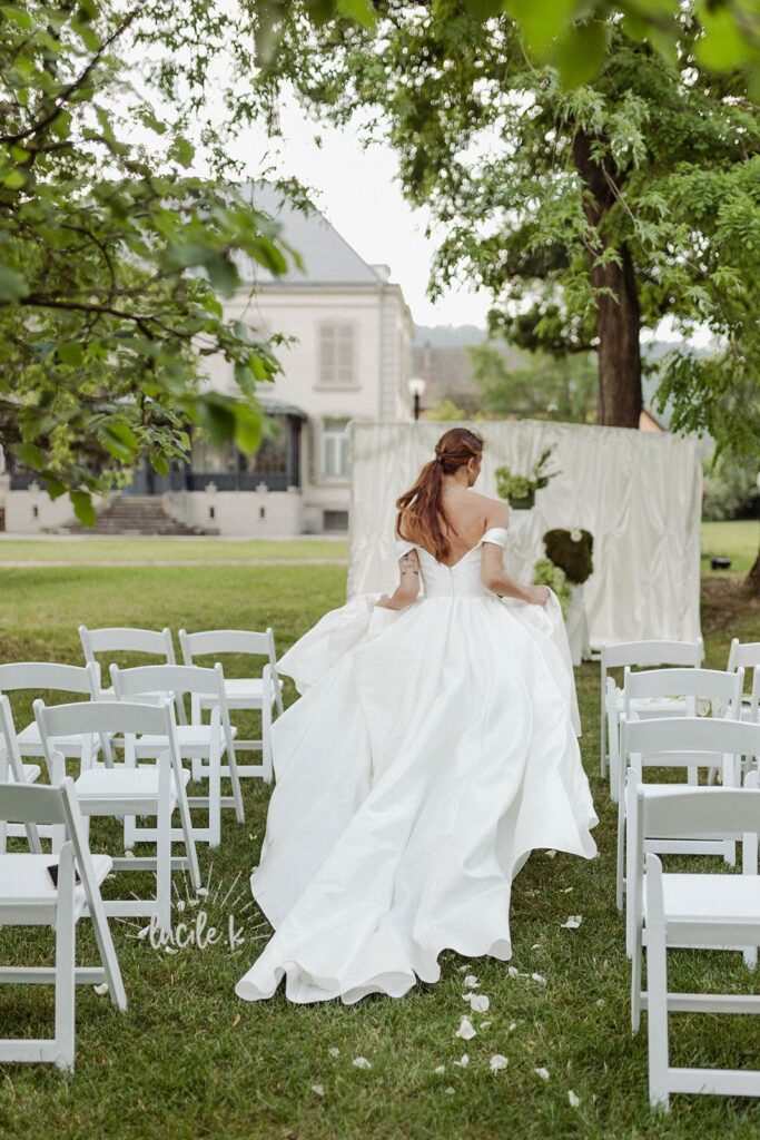 Shooting Editorial – La villa des hêtres pourpres Bride in a white wedding gown walking away down the aisle of an outdoor ceremony with white chairs and green trees surrounding her