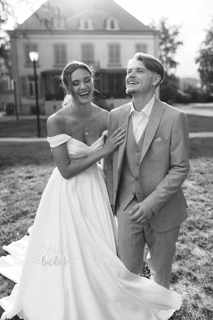 Shooting Editorial – La villa des hêtres pourpres Bride in an off-shoulder wedding gown with a long train laughing next to a groom in a light gray suit on a sunny lawn, a large building in the background.