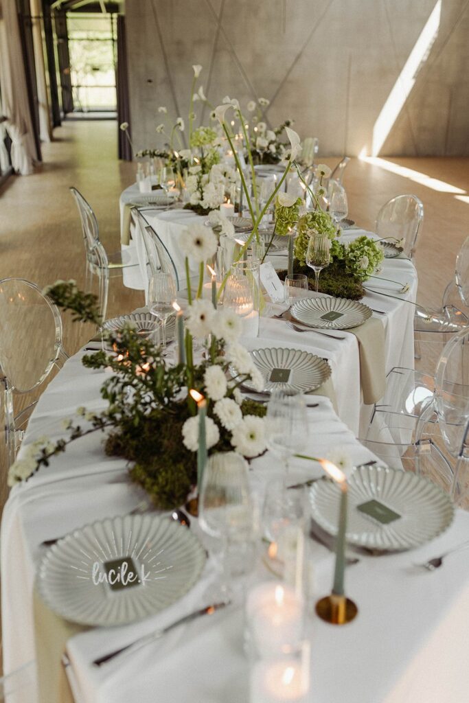Shooting Editorial – La villa des hêtres pourpres Long banquet table set for a formal event with white linens, clear chairs, and greenery-centered floral arrangements amid candlelight.