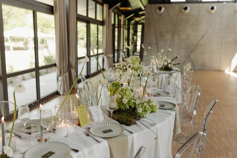 Elegant dining table set for a wedding with white linens, clear chairs, green-and-white floral centerpieces, and candles.